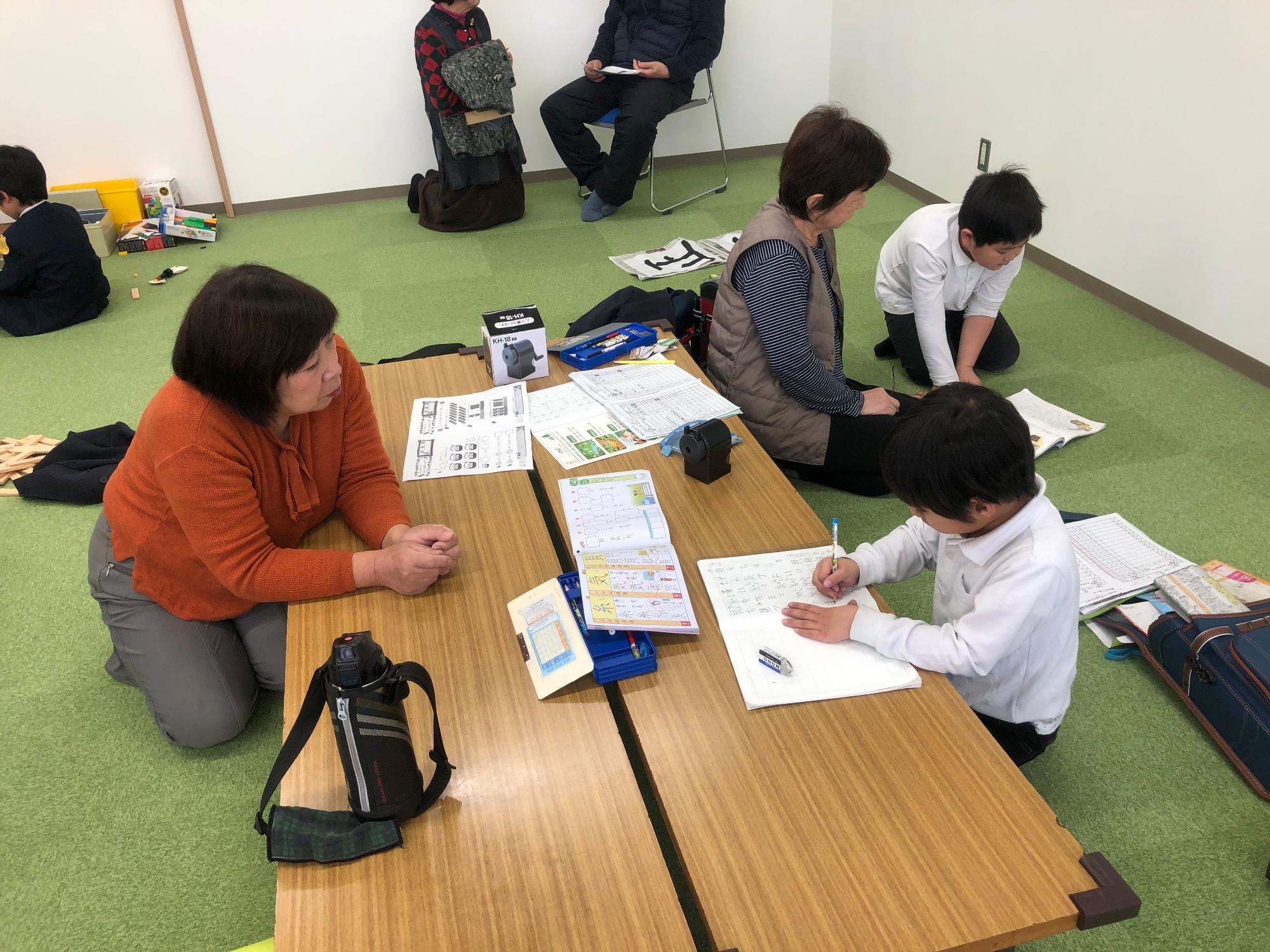 オレンジ色のセーターを着た女性の指導員が、机に向かってノートに書き物をしている男の子をすぐ横で見守っている写真