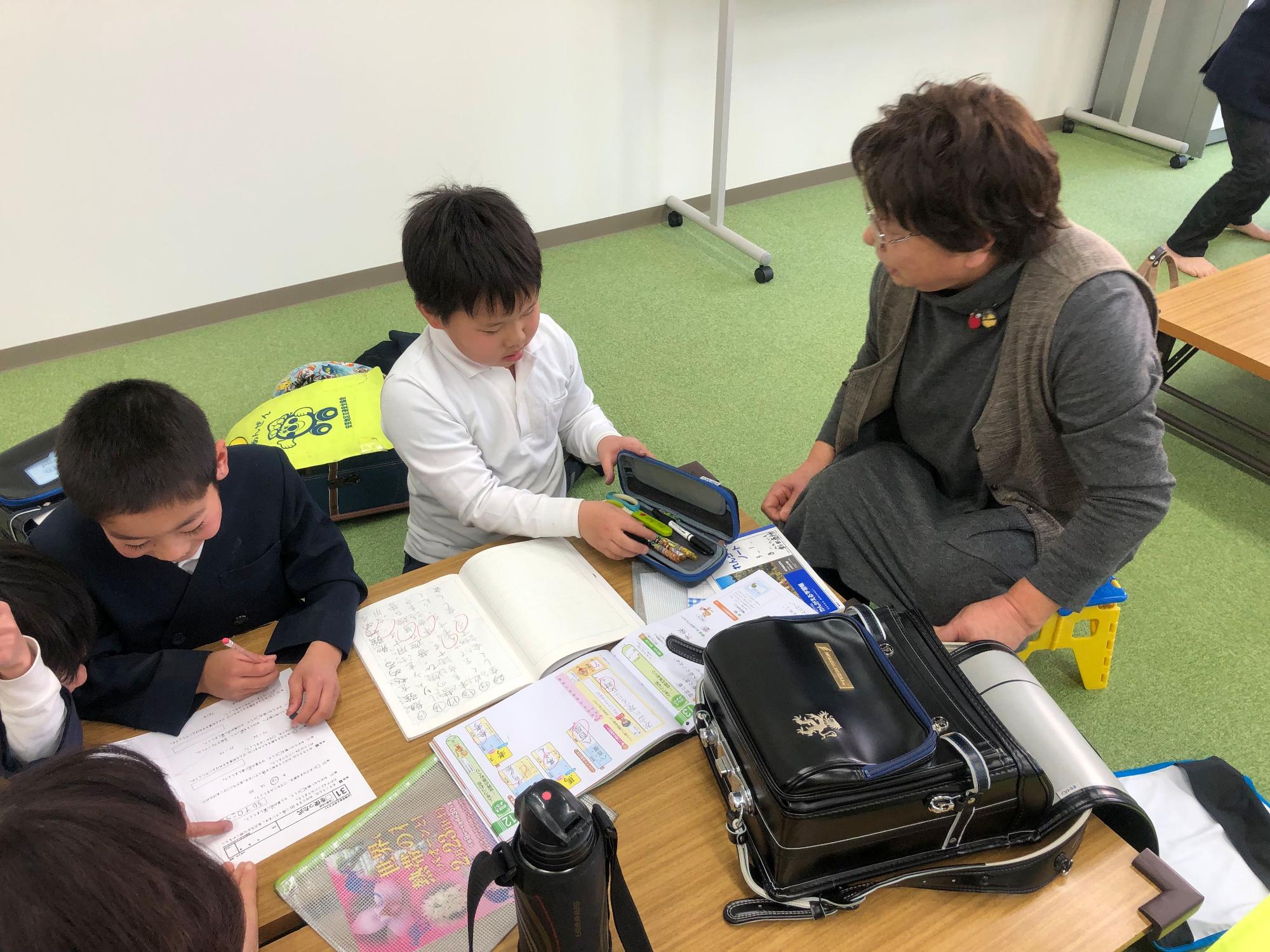 二人の男の子が机を並べてノートを広げて座り、そばで女性の指導員が見守っている写真