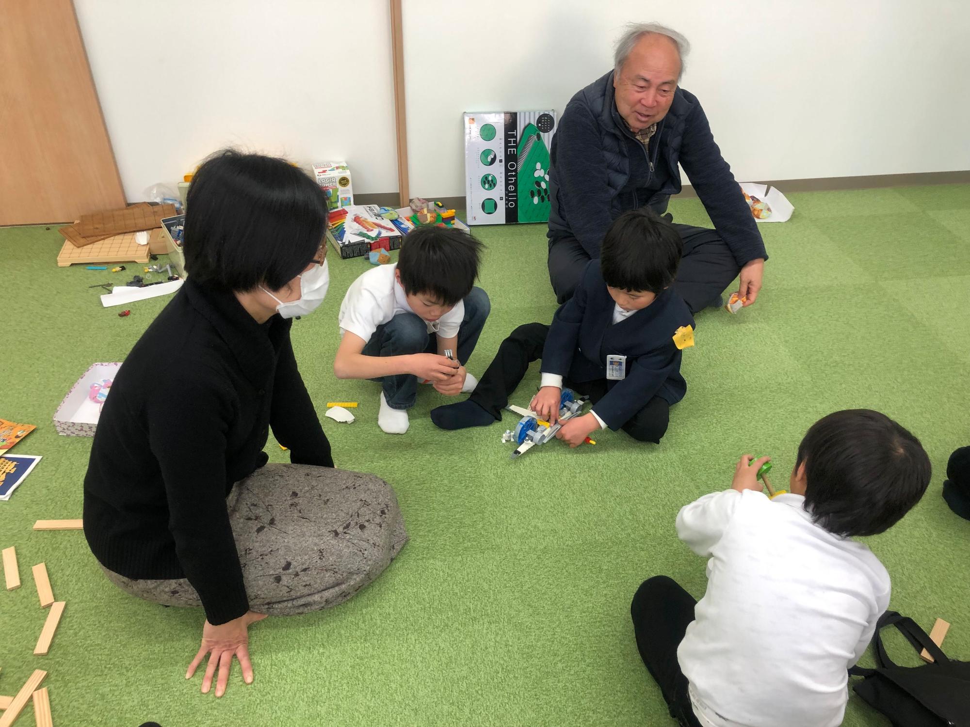 3人の男の子が元気に遊んでいる様子を、女性と年配の男性の指導員が少し離れた場所から優しく見守っている写真