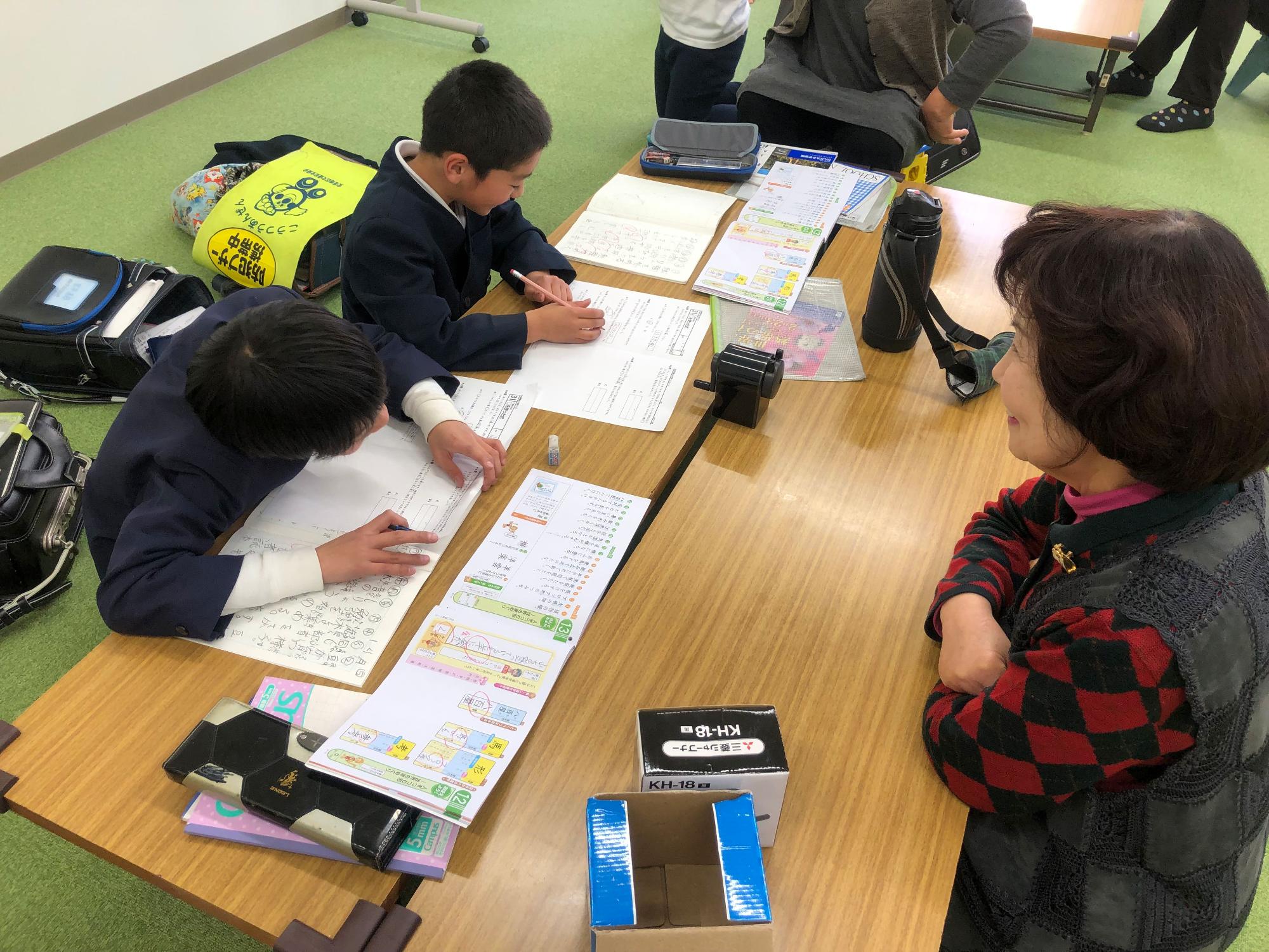 二人の男の子が机で並んでノートを広げて座り、前にベストをきた女性が見守っている写真