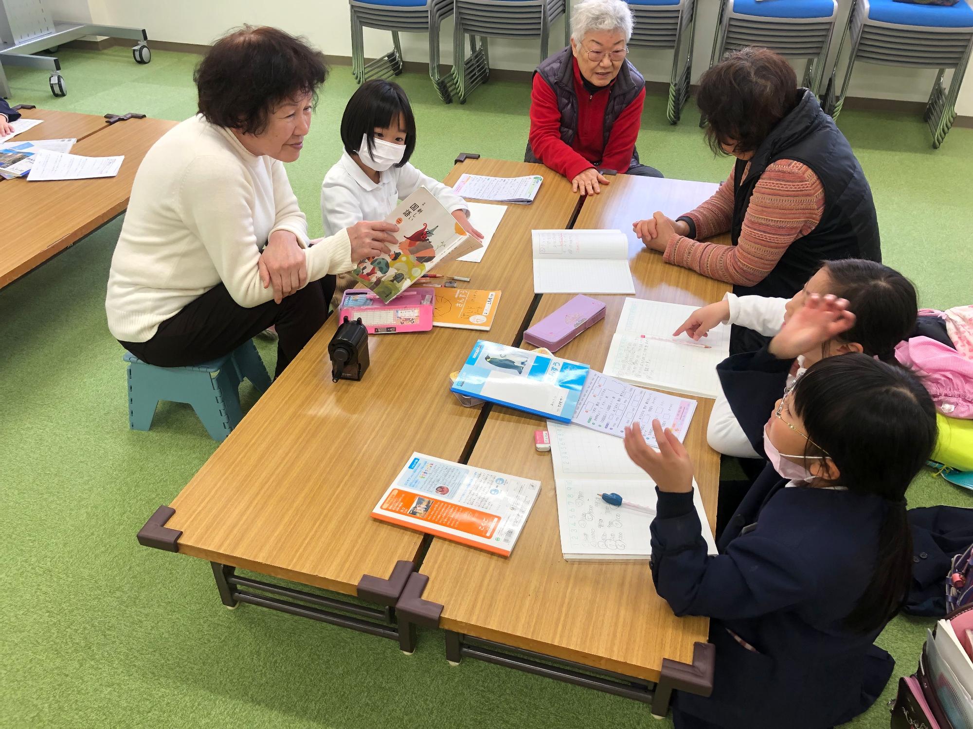 年配の女性指導員と子どもたちがテーブルを「コの字」に囲んで座っている写真