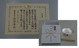左側に「お祝い状」があり、右下に銀杯と、赤い水引ののしがつけられた白い記念品が並んでいる写真