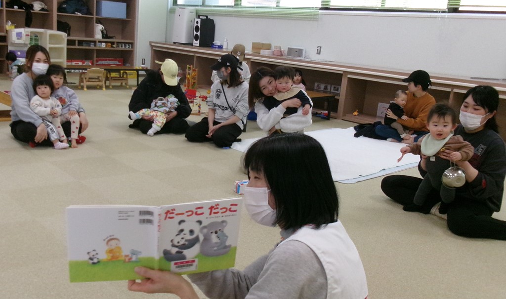 お母さんに抱っこしてもらいながら絵本を見ている子どもたち