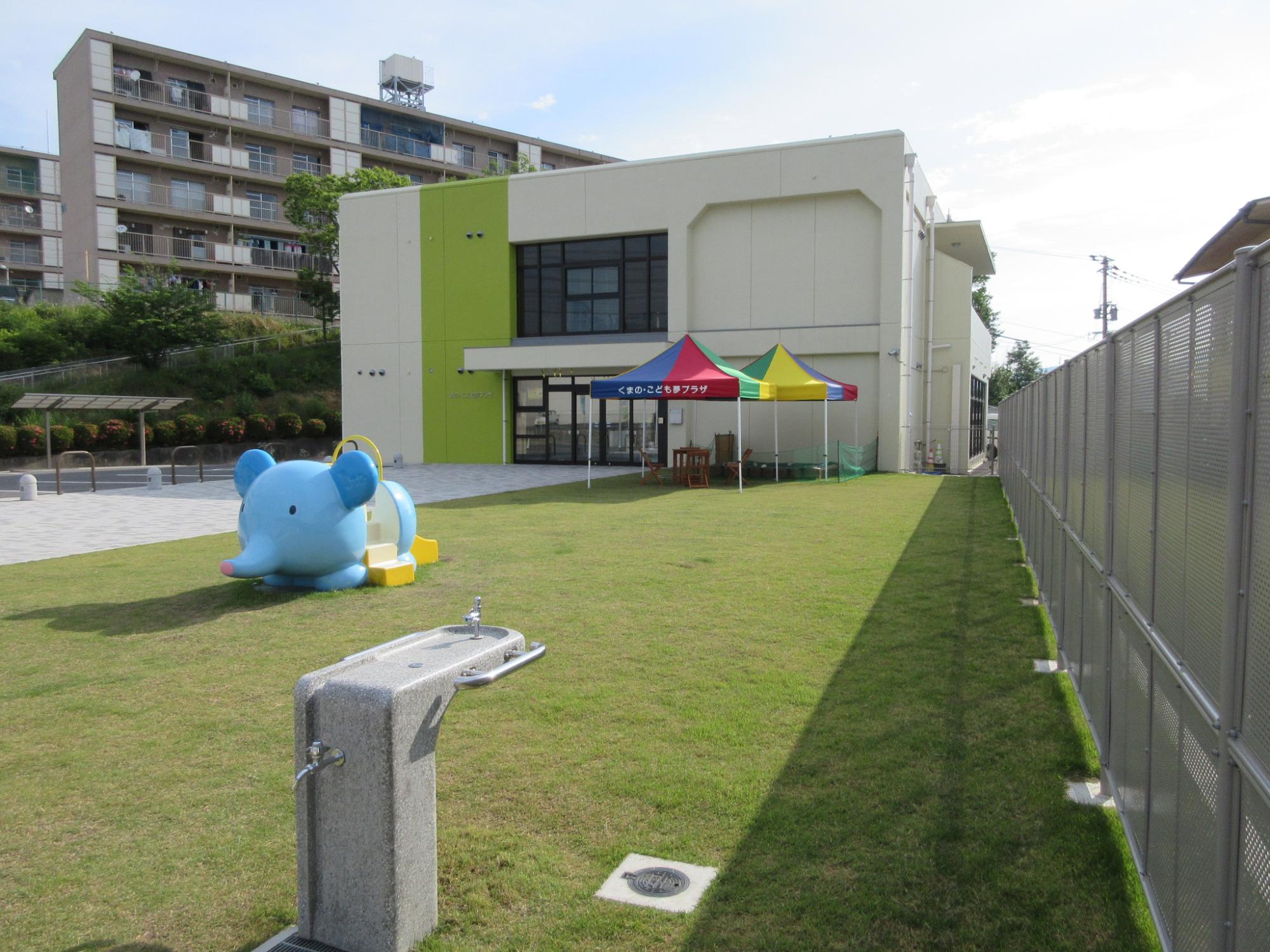 建物の前のスペースに芝生があり水飲み場とゾウの遊具がある写真