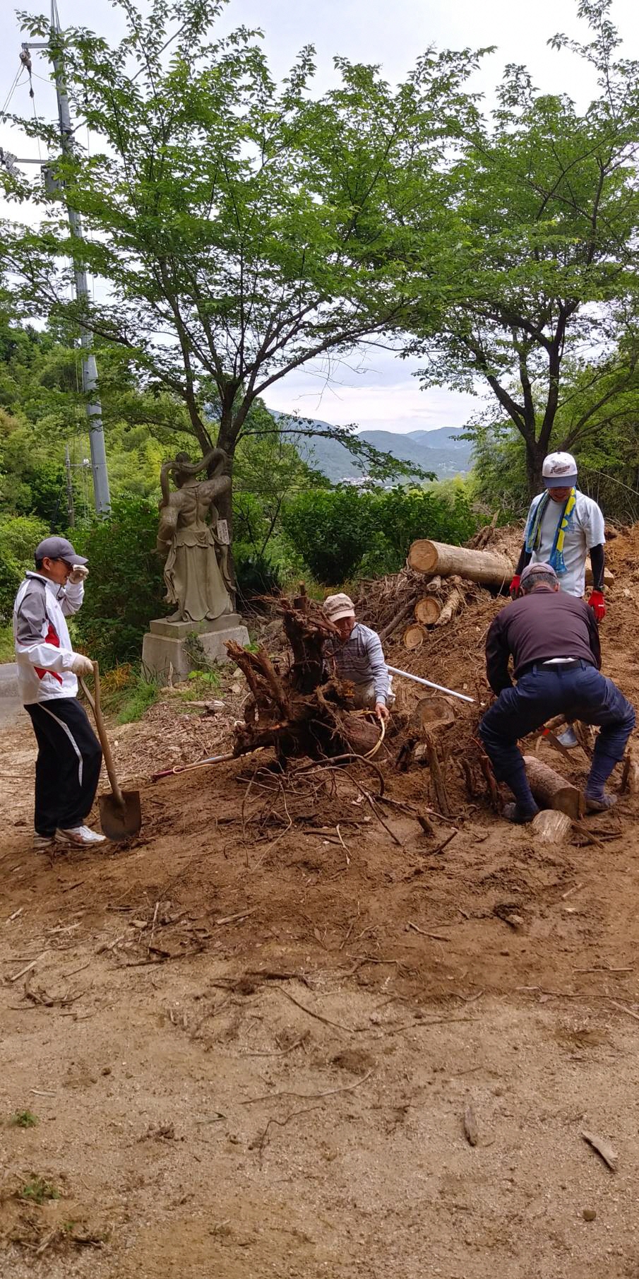 伐採された木や枝が並んでおり、シャベルやロープを使って4人の人物が作業をしている写真