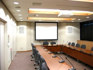 各席にマイクが設置されている会議用テーブルやスクリーンがある部屋の写真