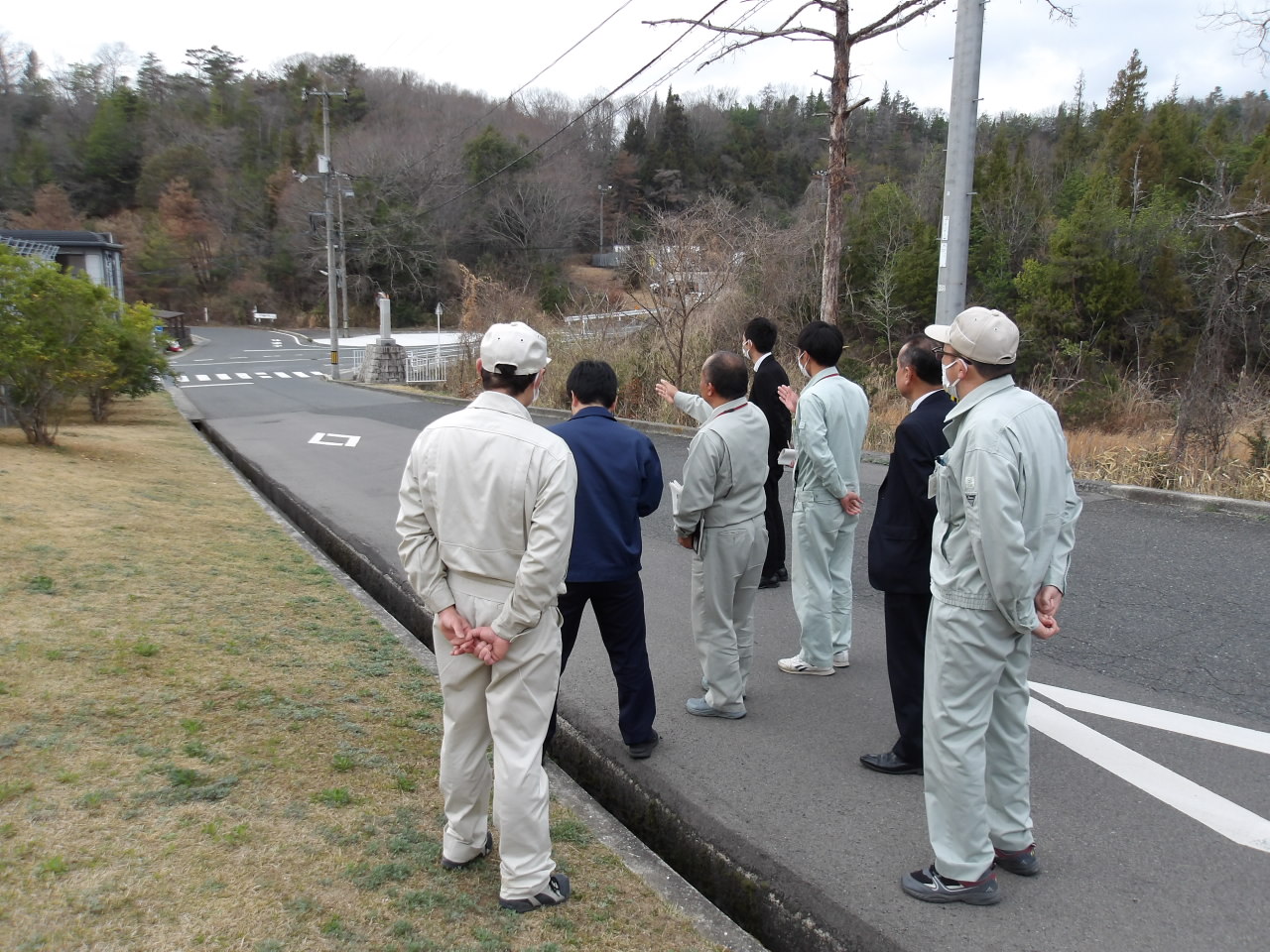 道路沿いでスーツ姿や作業服の参加者たちが現地の状況を確認している後ろ姿の写真