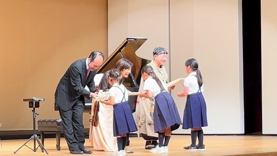 舞台上のピアノの前で子どもたちと握手をかわす三又治彦さん遠藤さつきさんの写真