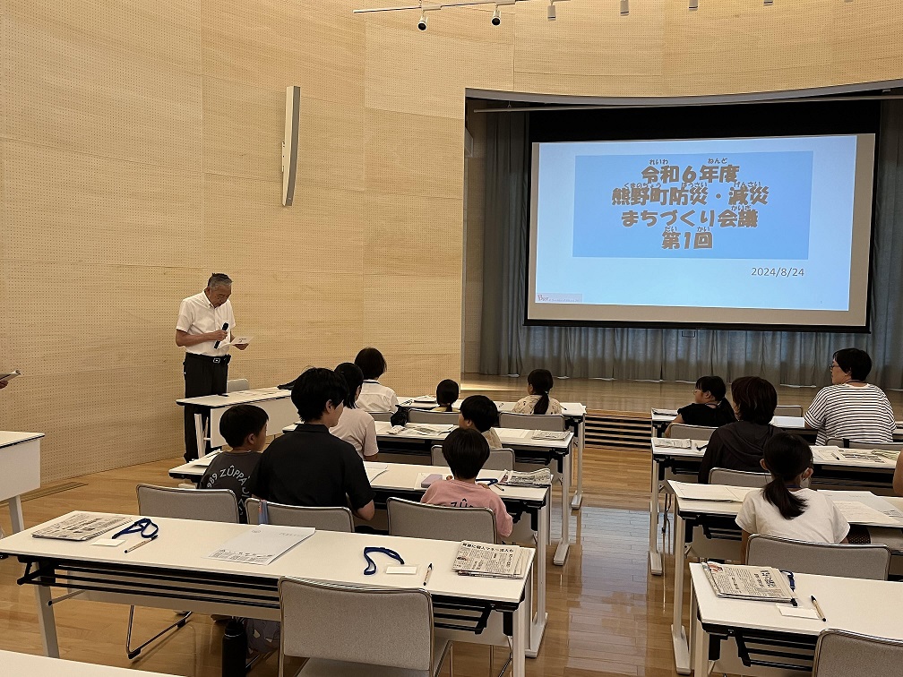 会議でマイクを持って話をする熊野町長とそれを聞いている親子の写真