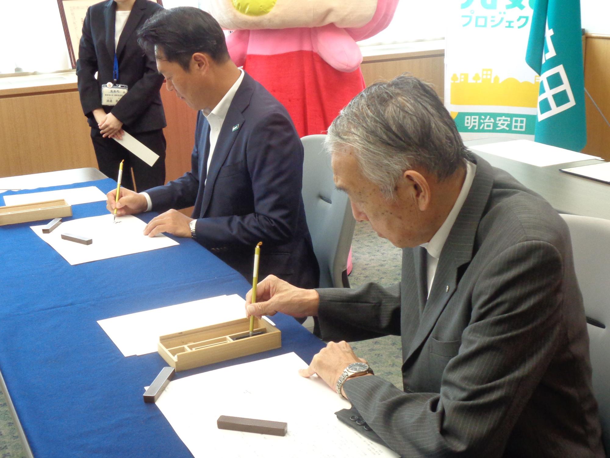 毛筆で半紙に文字を書こうとしている熊野町長と明治安田生命保険相互会社の担当者の写真