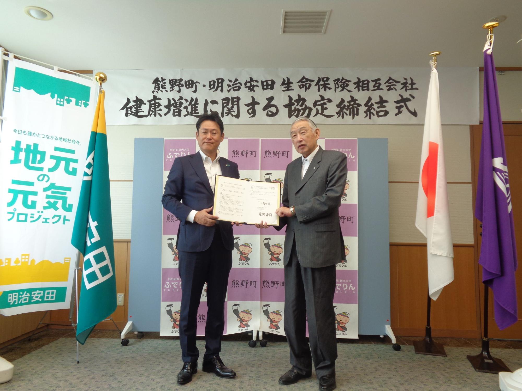 協定締結式で協定書を手に持ち記念撮影に臨む熊野町長と担当者の男性の写真