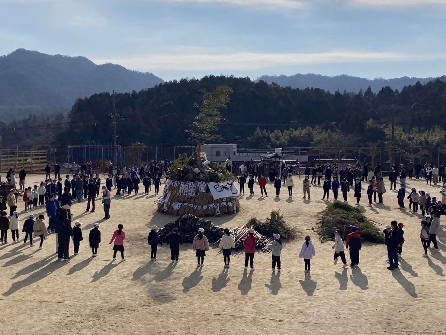 しめ縄や飾り物などを校庭の真ん中にまとめて周囲を子どもたちが取り囲んでいる様子を捉えた写真