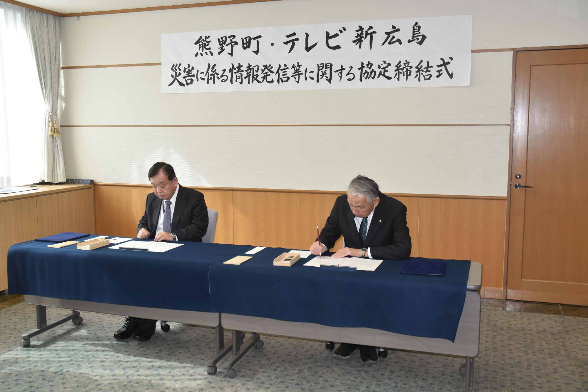 協定締結式で文書にサインする熊野町長とテレビ新広島の代表の写真