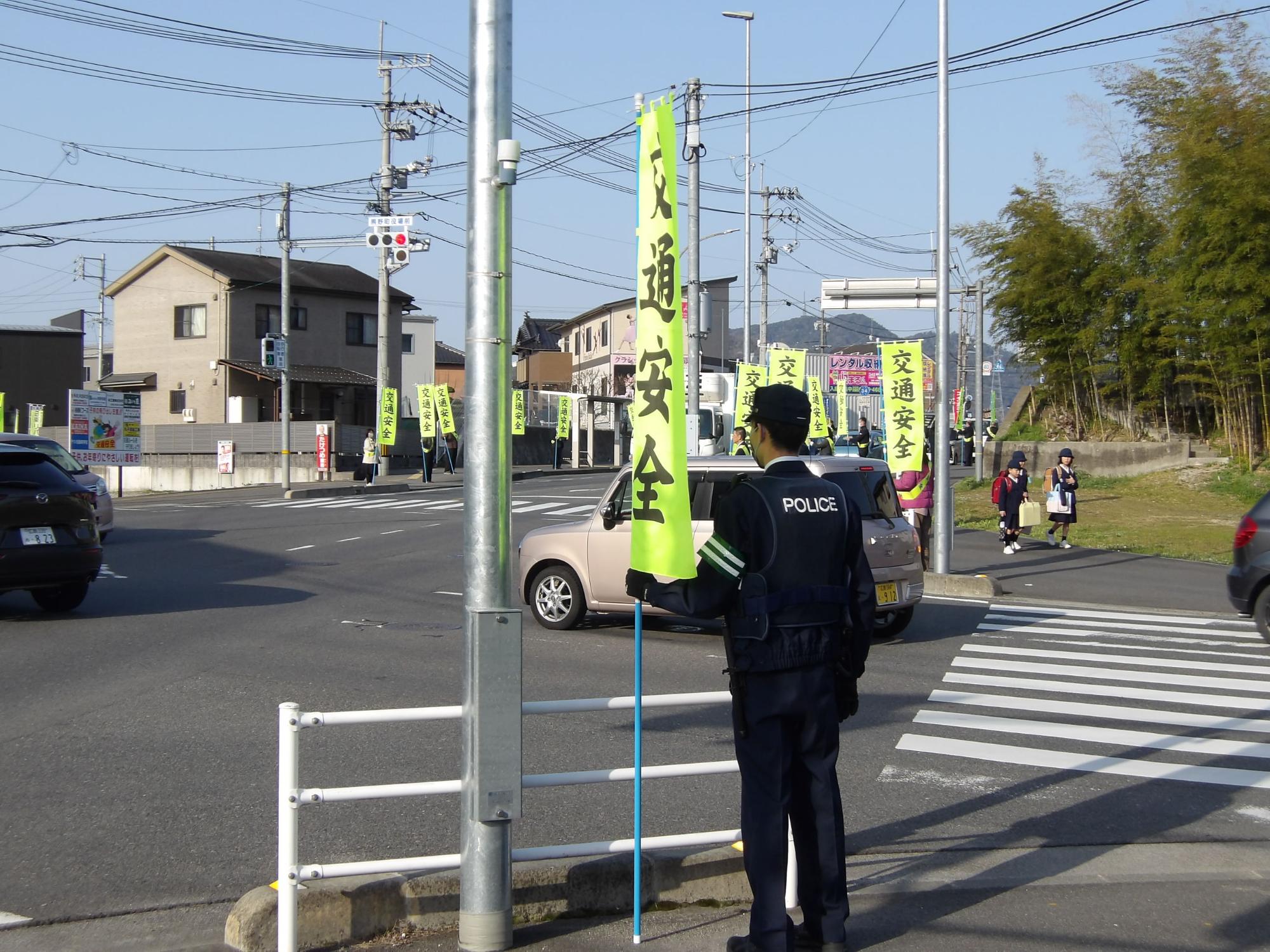 交通安全ののぼりを持ち、横断歩道で小学生の登校を見守る警察官の写真