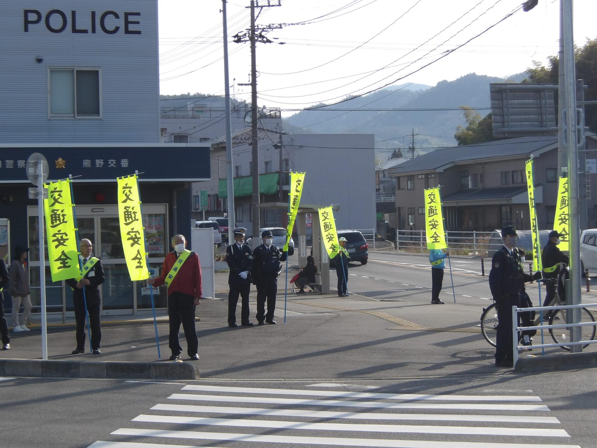 交番前の横断歩道で交通安全ののぼりを持ちキャンペーンを行う警察官や地域住民の方々の写真