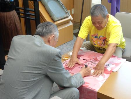 黄色のティーシャツを着ている人が持ってきたピンク色の旗のようなものにコメントを記入している熊野町長の写真