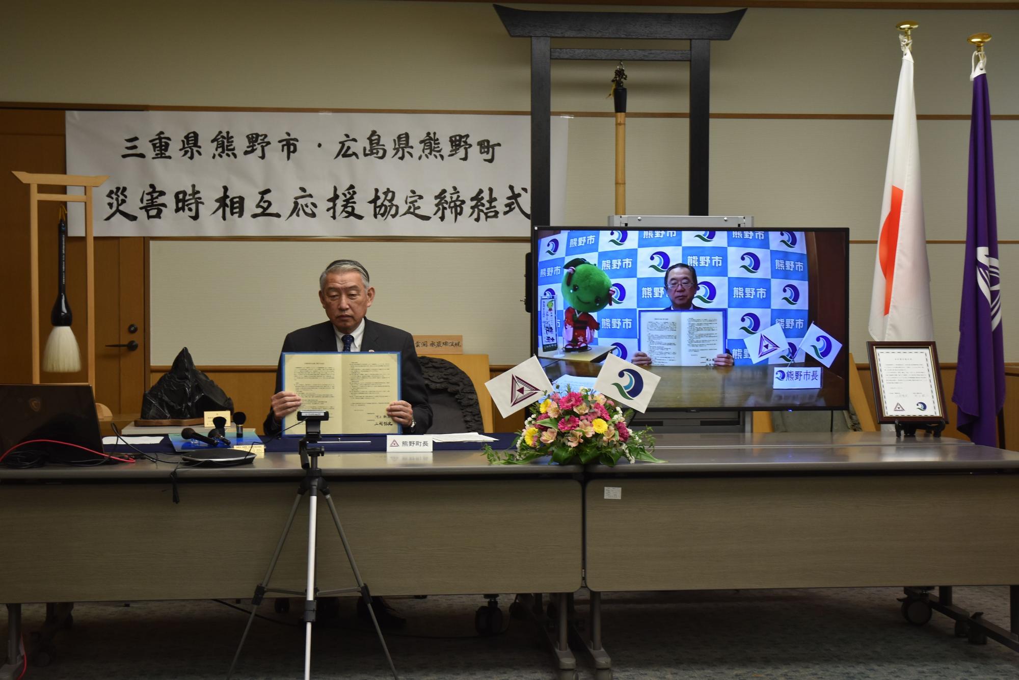 三村町長とモニターに映っている河上市長が協定書を広げて見せている写真