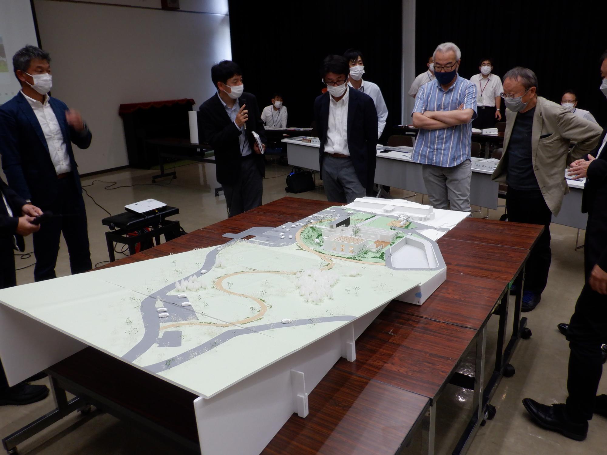 公開ヒアリングでテーブル上のジオラマの説明を受けている有識者の写真