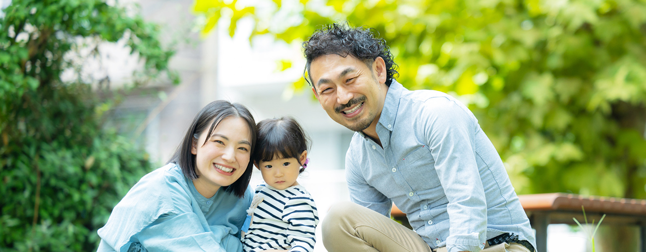 新緑の屋外で、笑顔の両親がボーダー柄の服を着た幼い子供を挟んで並び、カメラに向かって微笑んでいる様子