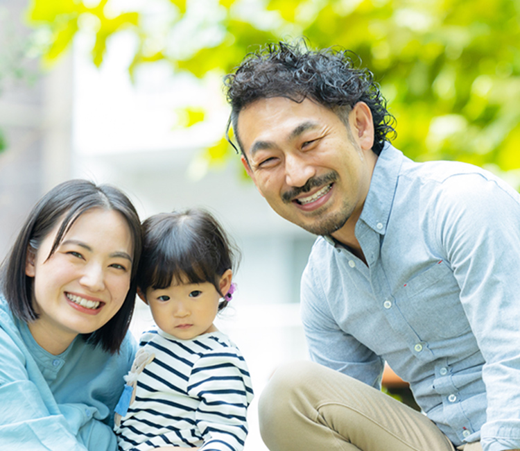 新緑の屋外で、笑顔の両親がボーダー柄の服を着た幼い子供を挟んで並び、カメラに向かって微笑んでいる様子