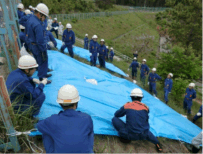 野外の坂にブルーシートが敷かれた周りに消防団の人たちが集まっている写真