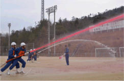 広いグラウンドで消防団の人たちが消防ホースで放水している写真