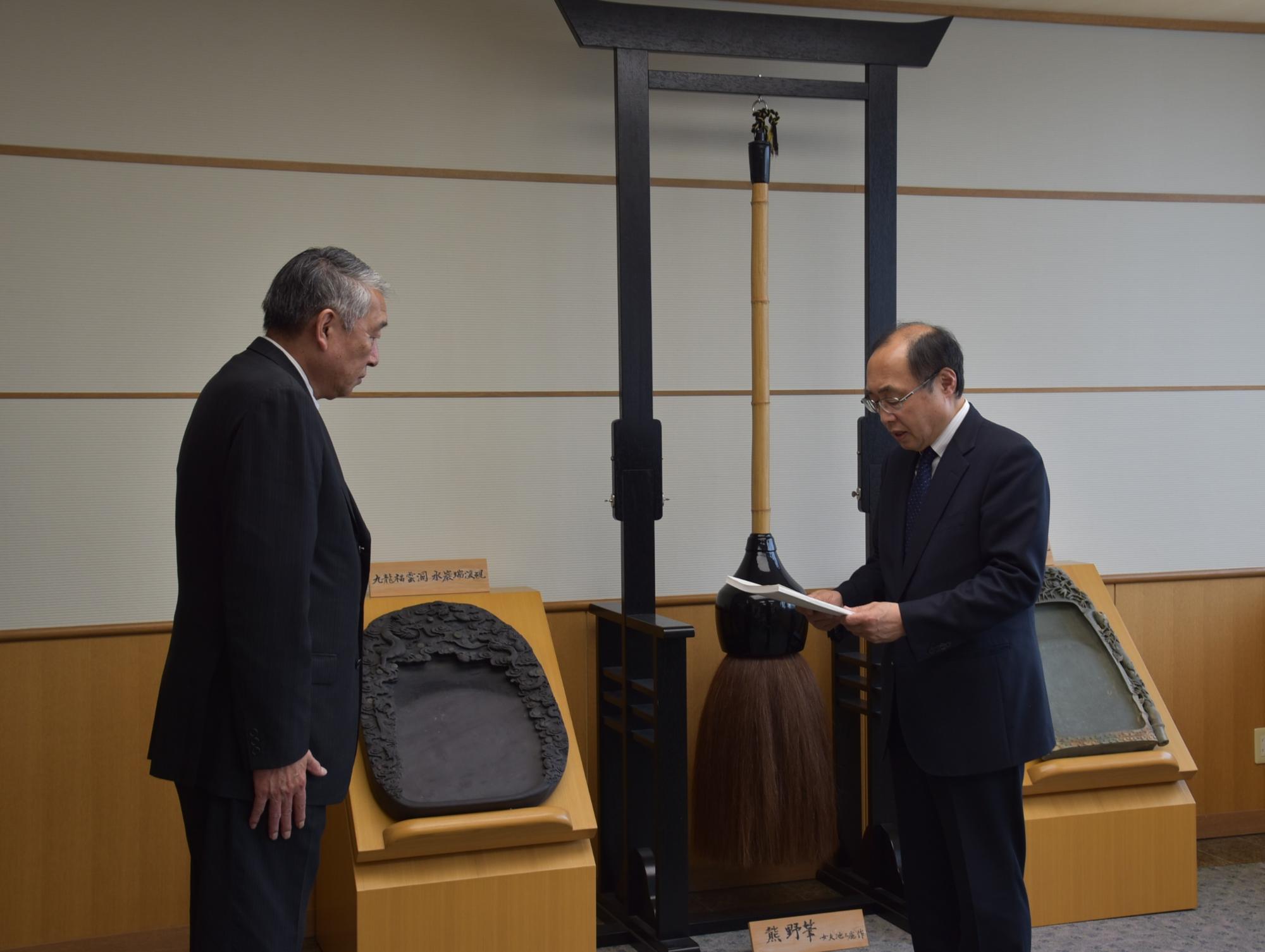 男性が書類を両手に持ち読んでいる向かい側で男性が起立している写真