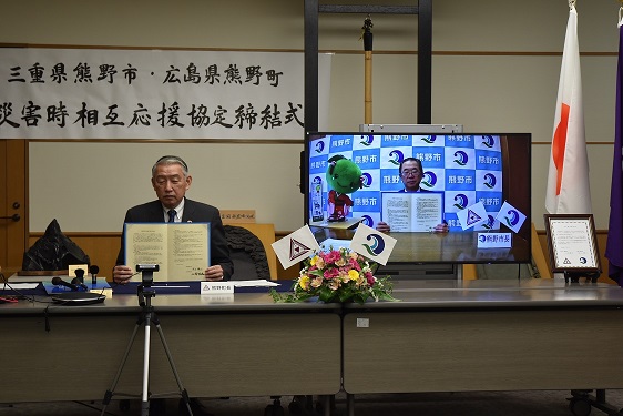 災害時の相互応援協定締結式で、男性が協定書を掲げており、その右においてあるモニターには協定書を掲げた別の男性が移っている写真