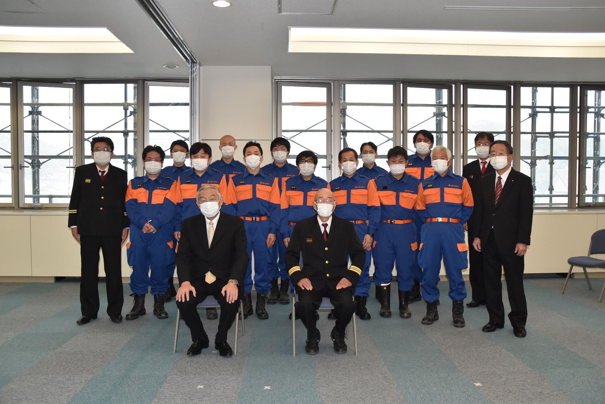 男性二人が椅子に座っておりその後ろに消防団の人たちが横二列に並んでいる写真