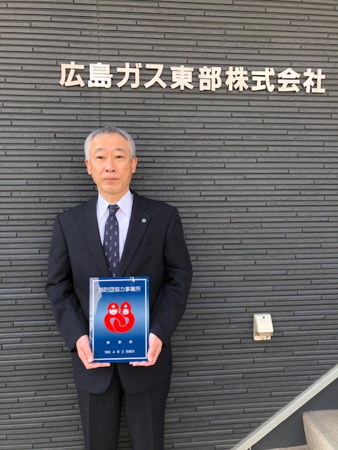スーツ姿の男性が「広島ガス東部株式会社」と書かれた壁の前で消防団協力事業所の認定証を持っている写真