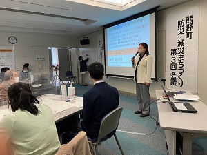 画像が移されたスクリーンの横でマイクを持って話をしている人の写真