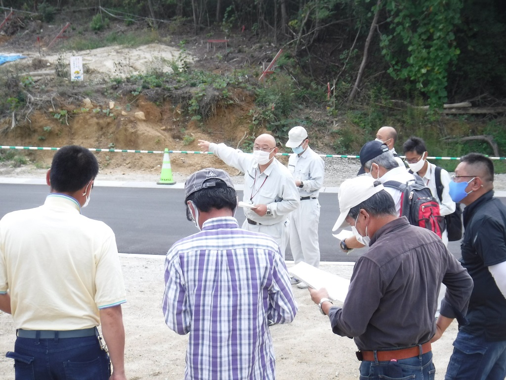 崩れて赤茶色の土が見えている道路脇の山肌を指差す男性と、それに付き従う男性達の写真