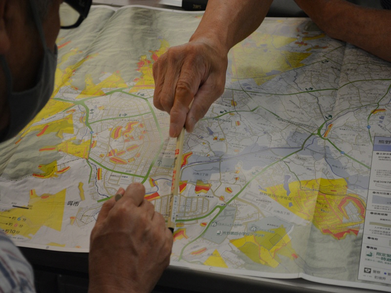 黄色やオレンジ色で塗り分けされた地図を広げ、ペンで地図を指し示しながら話し合いを行う人達の写真