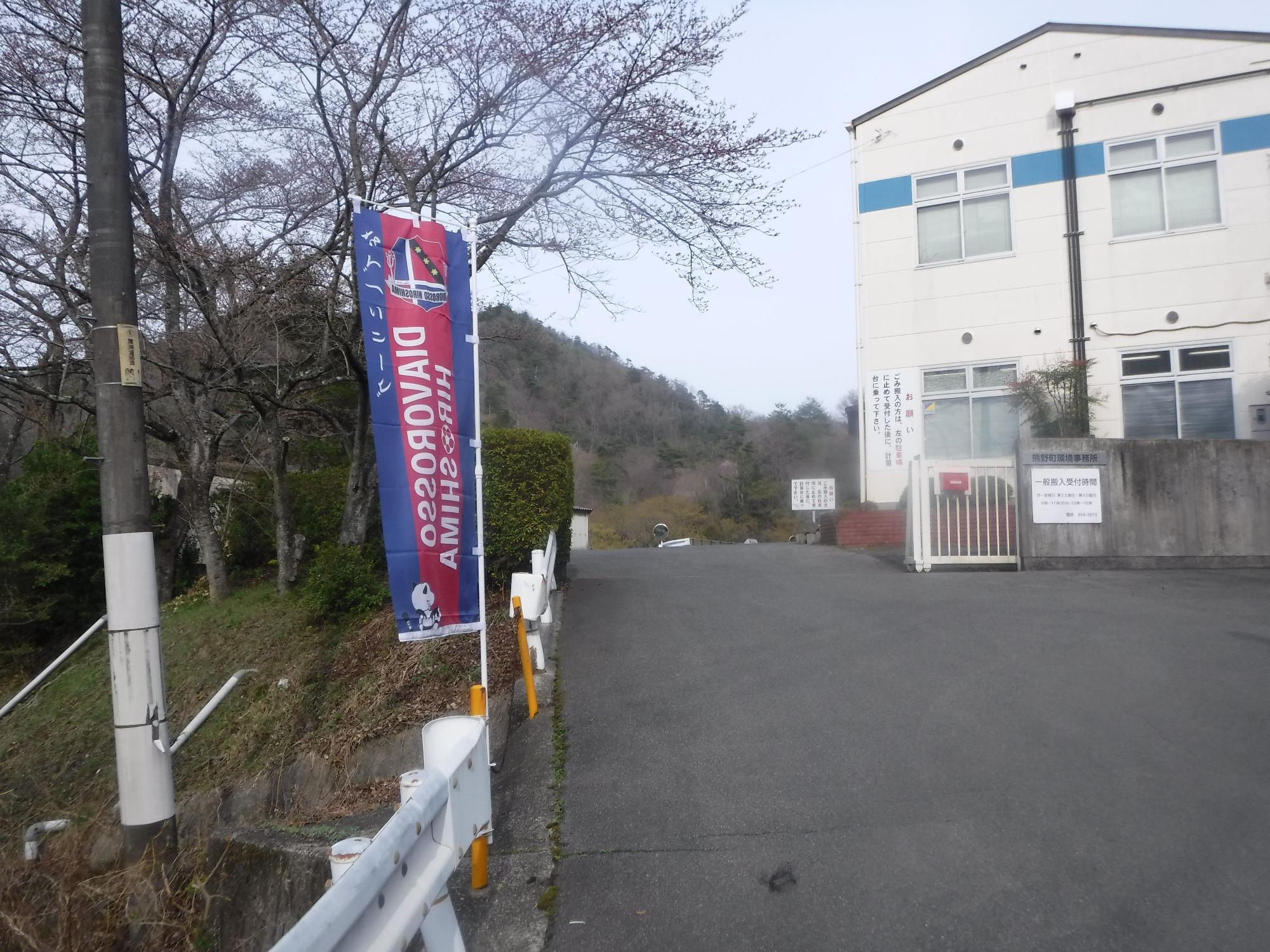 建物へ続く坂道の入口付近に門があり、そばにのぼり旗が立っている写真