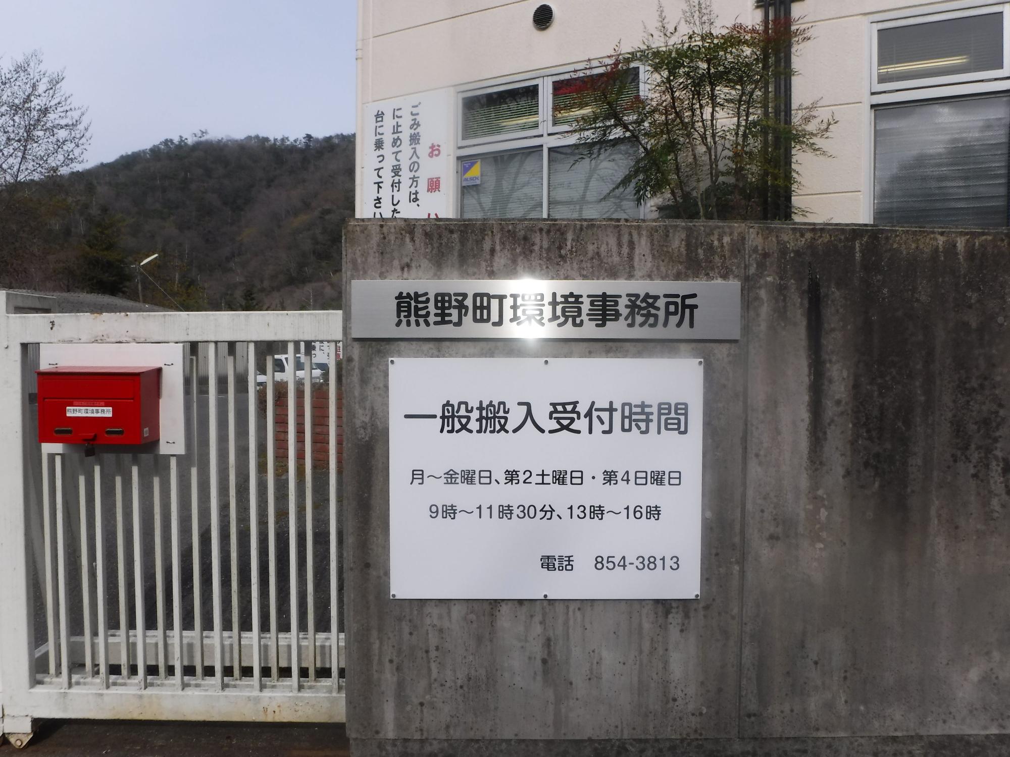建物の入口付近の壁に、環境事務所の名称と一般搬入受付時間が書かれた案内板が設置されている写真