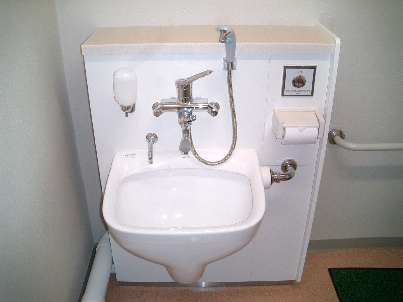 白の流し台があり、その横には蛇口やシャワー型の水栓、トイレットペーパーホルダーなどが備え付けられた写真