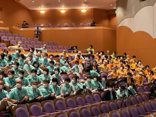 文化会館で黄色の洋服と薄緑色の洋服を着た人たちがリハビリ体操を行っている写真