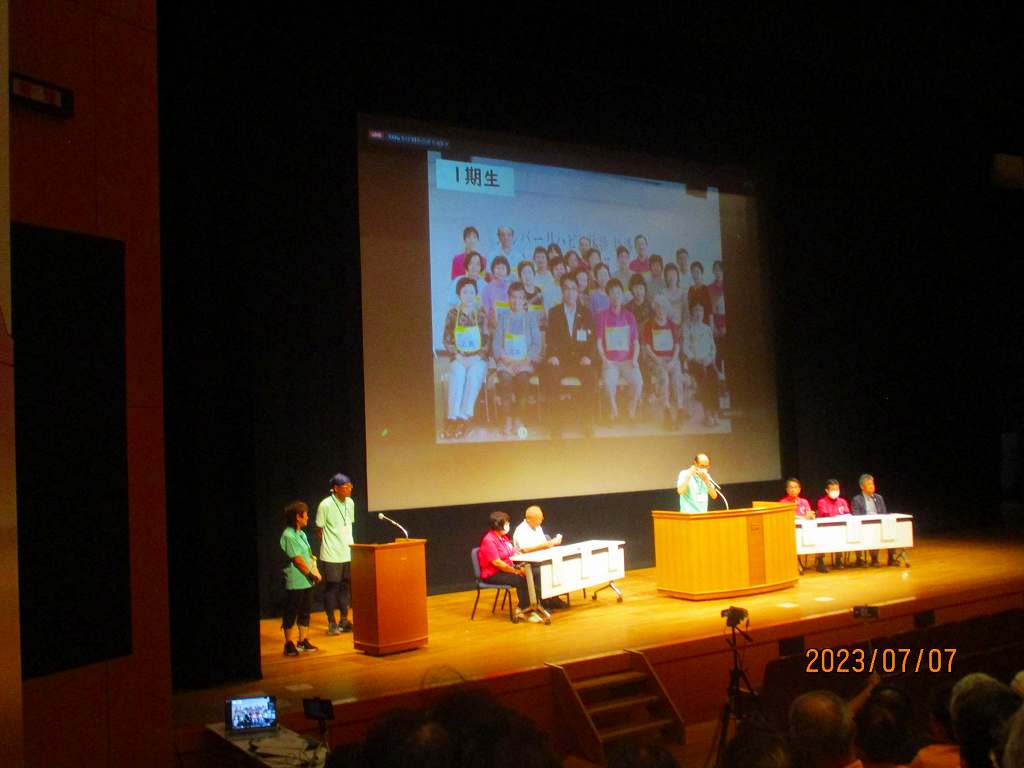 会館でスクリーンを用いて登壇した人たちが発表を行っている写真