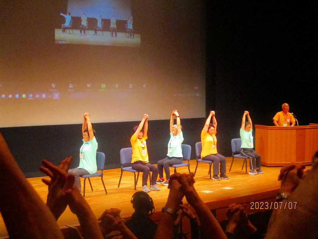 会館で椅子に座った人たちが体操を行っている写真