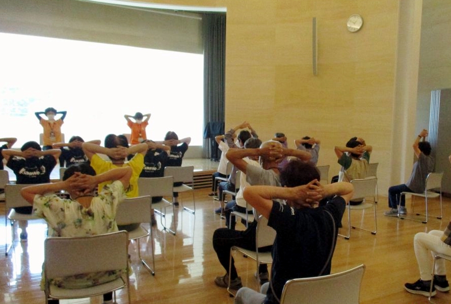 室内で椅子に座った人たちが後ろに手を組み体操を行っている写真