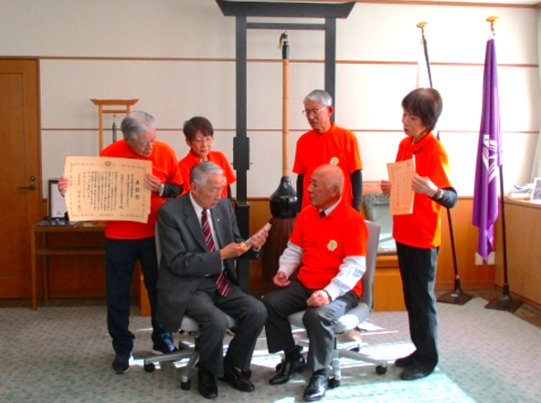 室内で赤い洋服の人たちが椅子に座った男性に注目している写真