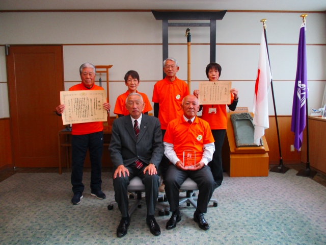 室内で赤い洋服の人たちが表彰状を掲げて記念撮影を行っている写真