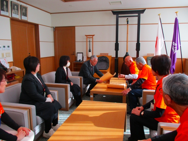 室内で椅子に座った人が表彰状を読みあげている写真