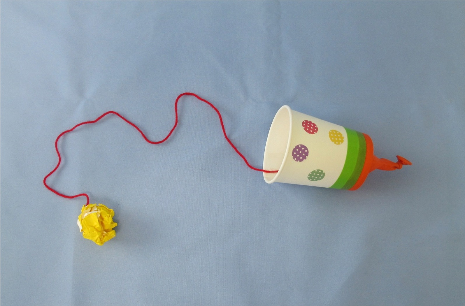 水色の背景の上で、紙コップに通した赤いひもが黄色の丸めた紙につながって置かれている糸電話の写真