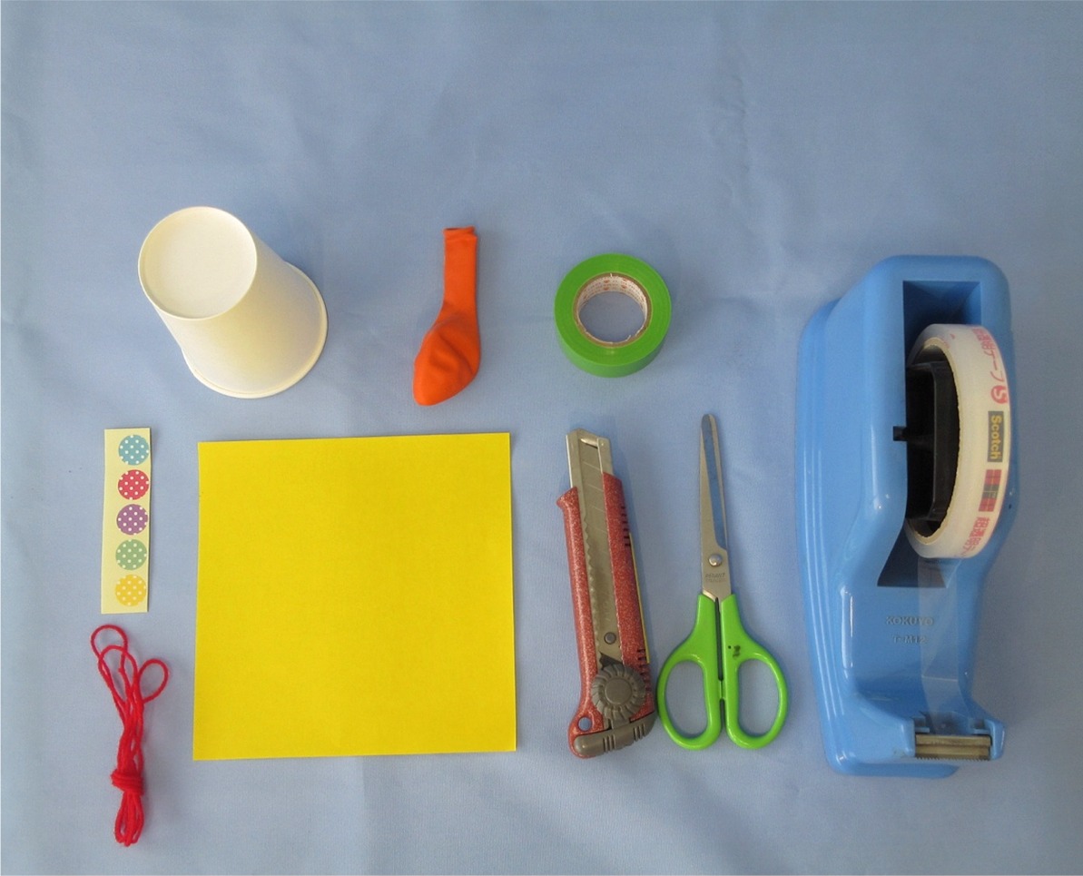 赤いひも、紙コップ、黄色い紙、カッターやハサミ、セロテープなどが並んで置かれている写真