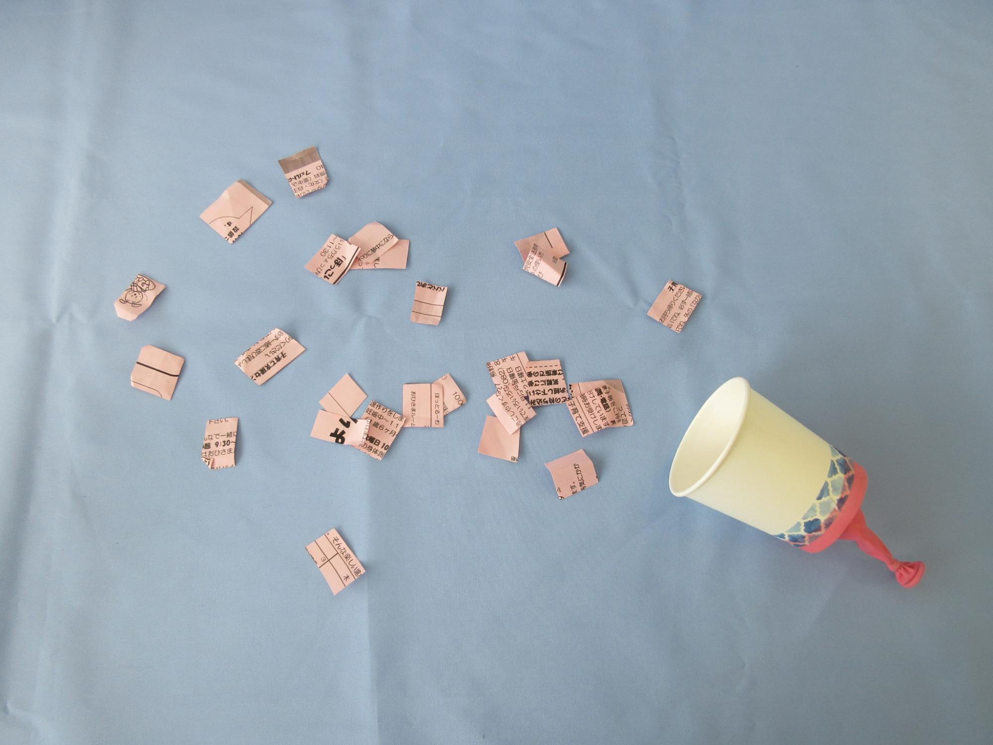 ピンク色のゴムが付いた紙コップのそばで四角形に刻まれた紙が散らばっている写真