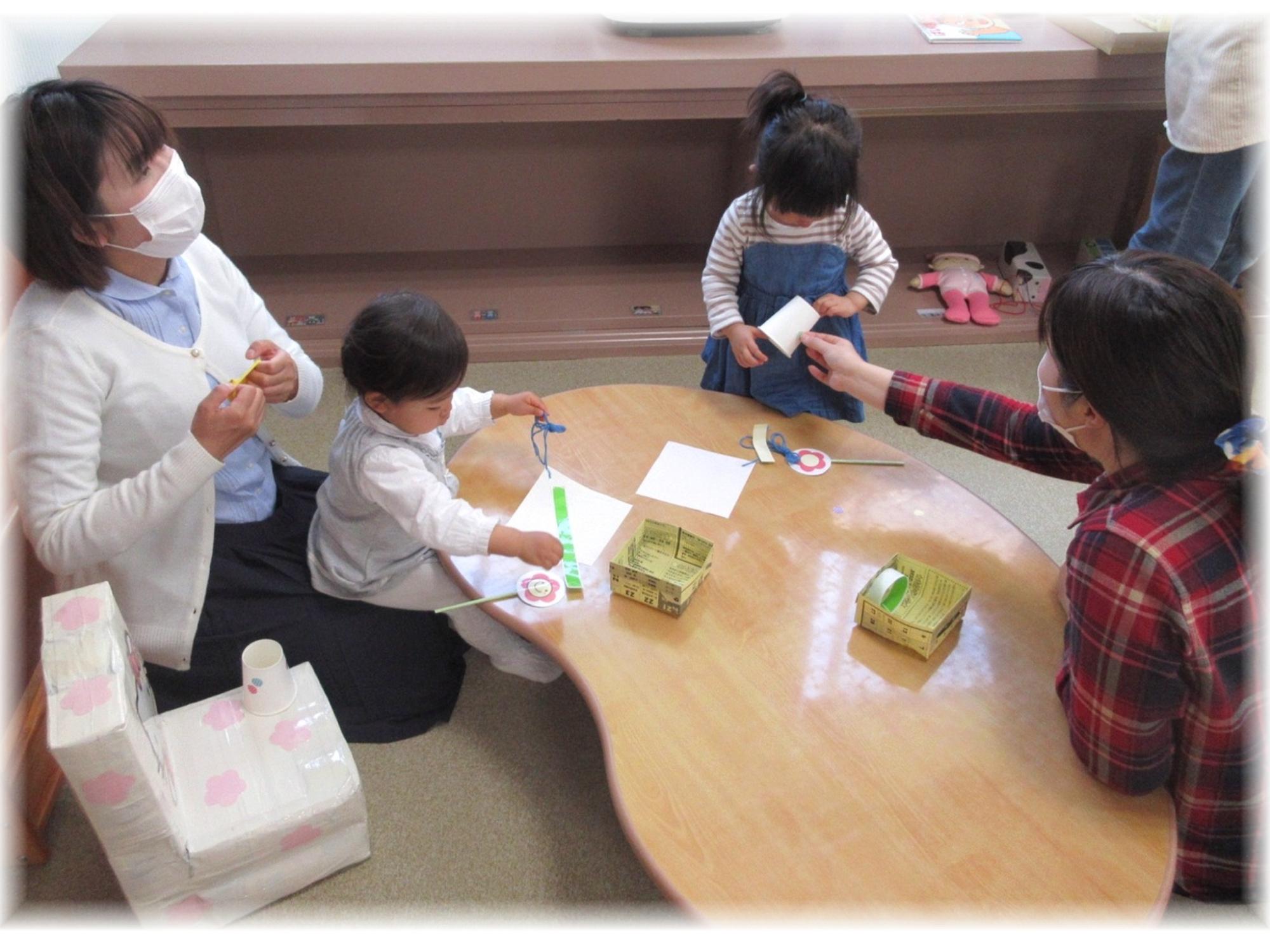 室内のテーブルを囲んで、大人に見守られながら幼い子どもたちが工作をしている写真