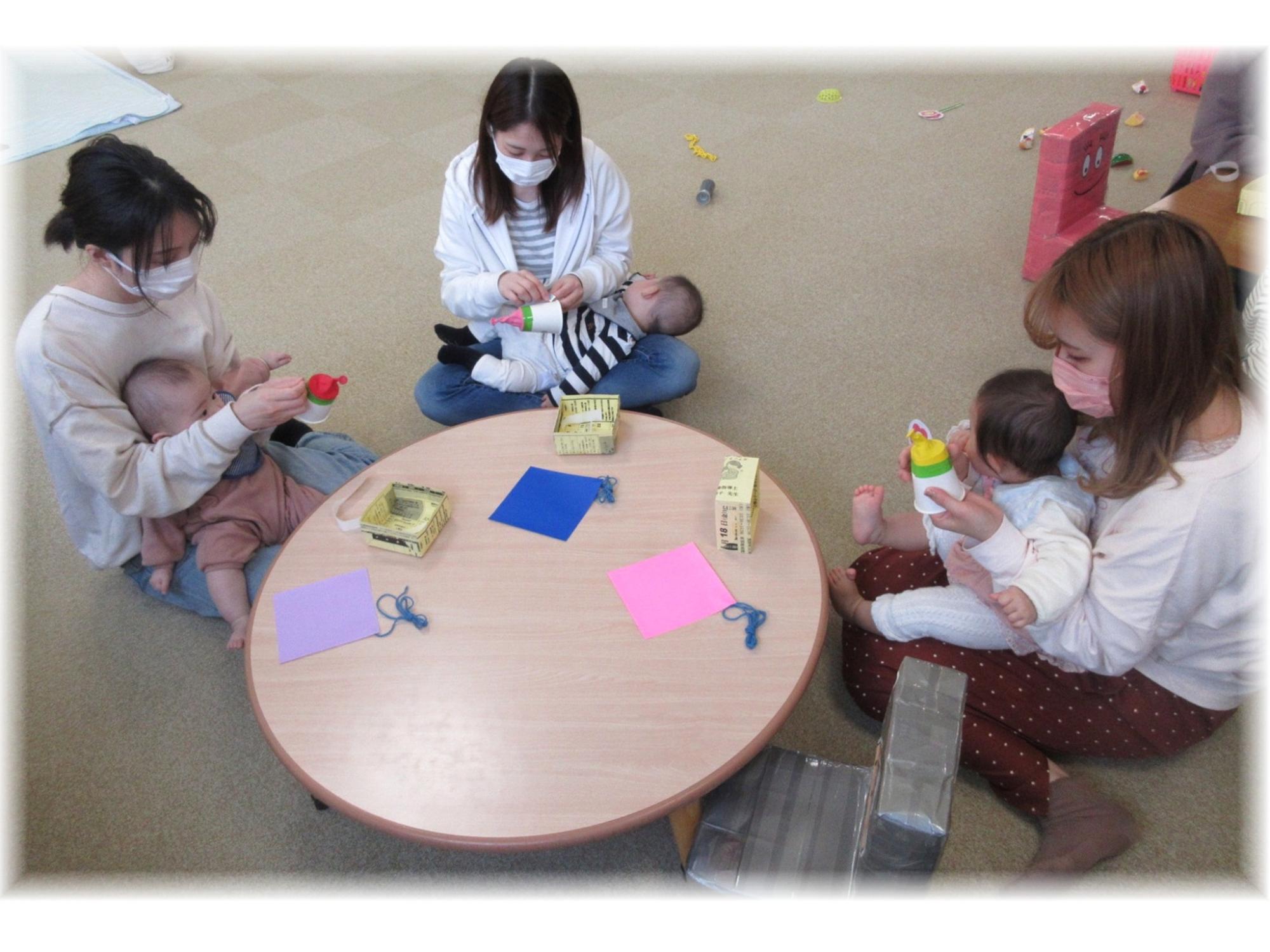 室内の丸いテーブルを囲んで、保護者が抱っこした乳児と一緒に紙コップを使った工作をしている様子の写真