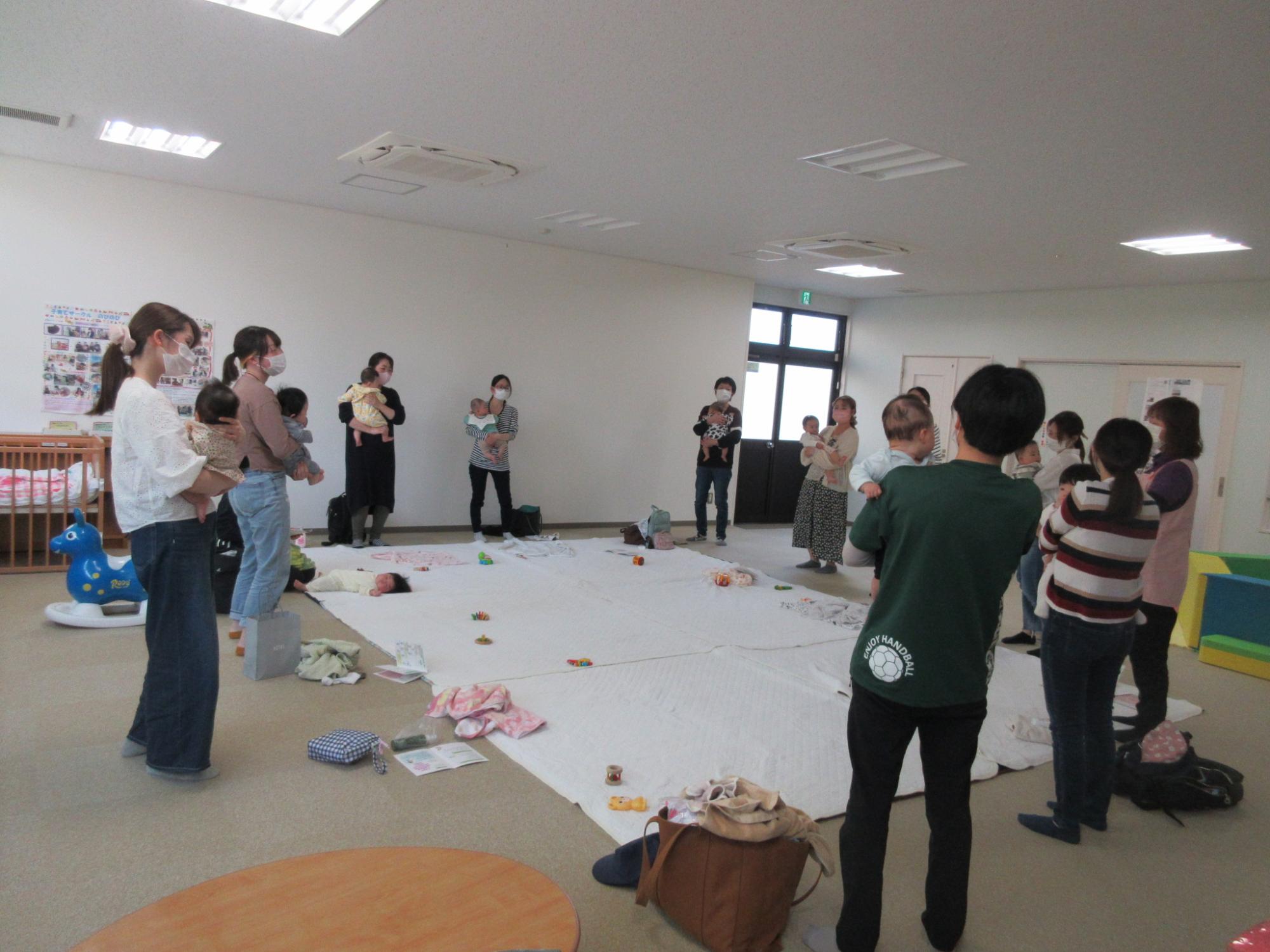 立ちながら赤ちゃんを抱いてマットを囲む女性たちの写真