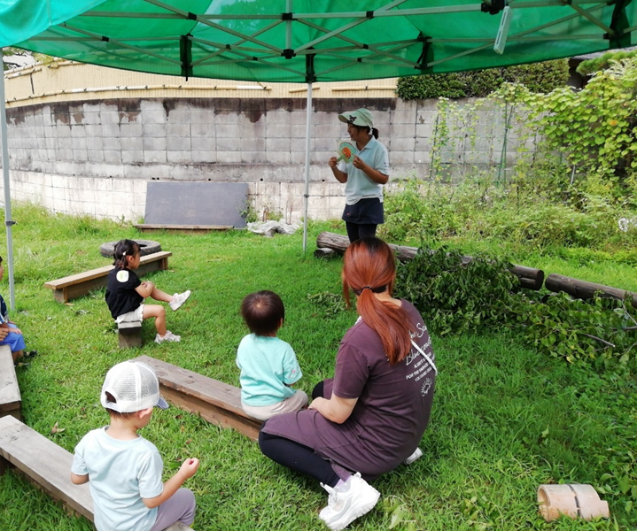 緑色をしたテントの屋根下で話を聞いている親子たちの写真